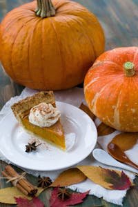 Slice of pumpkin pie with whipped cream, surrounded by pumpkins and autumn leaves, perfect for fall celebrations.