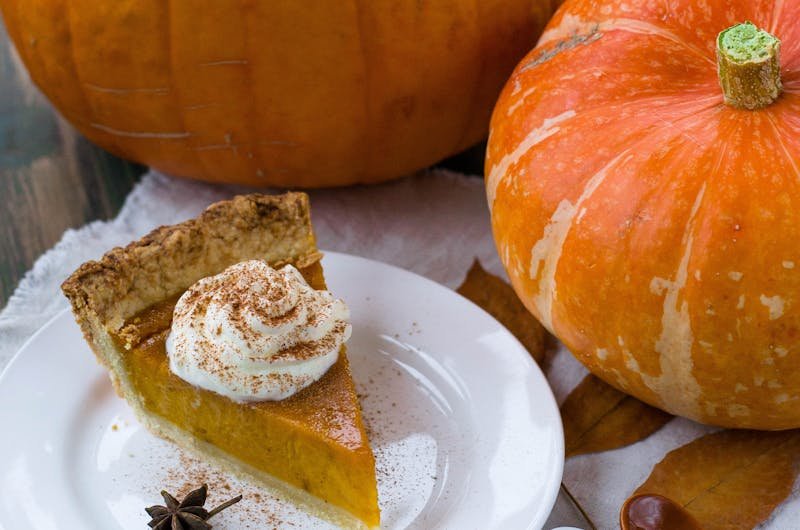 Slice of pumpkin pie with whipped cream, surrounded by pumpkins and autumn leaves, perfect for fall celebrations.